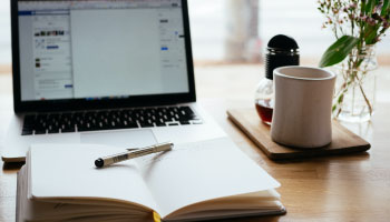 Close up of various items on a desk. Items include a mug, a journal, and a laptop.