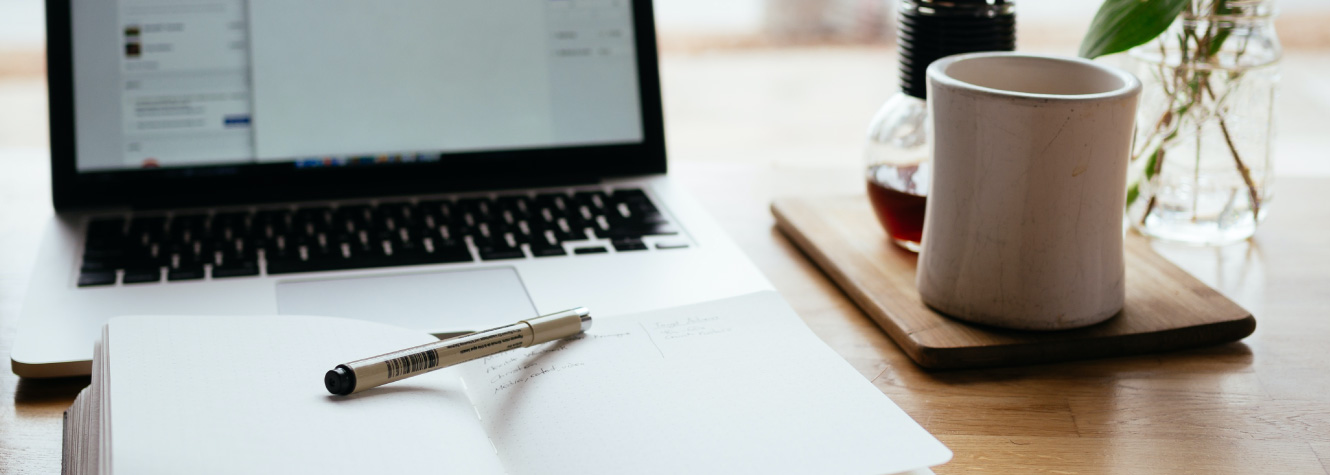 Close up of items on a desk, including a mug, journal, and laptop.