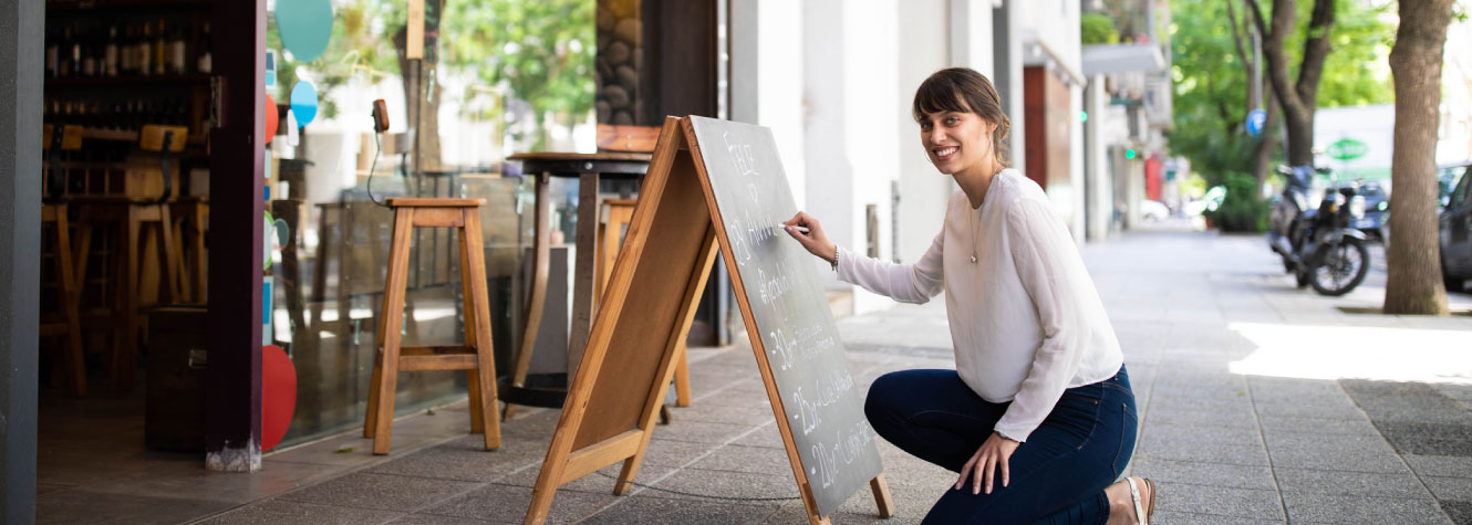 A young woman kneeling next to a chalk board outside of a retail store.