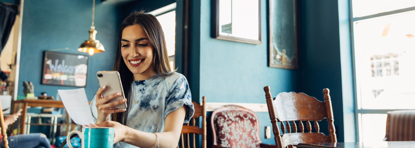 A young woman sitting in a cafe. She is holding a cell phone and a receipt.