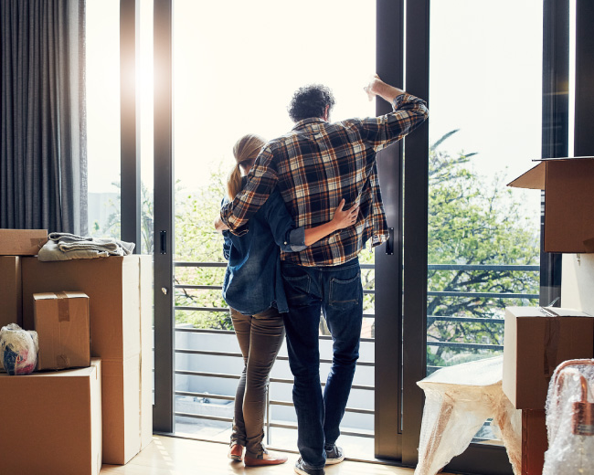 A young couple looking out a large window. There are cardboard boxes all around them.