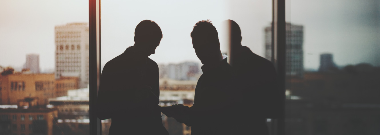 Two men standing in an office building with a large window and city skyline behind them.