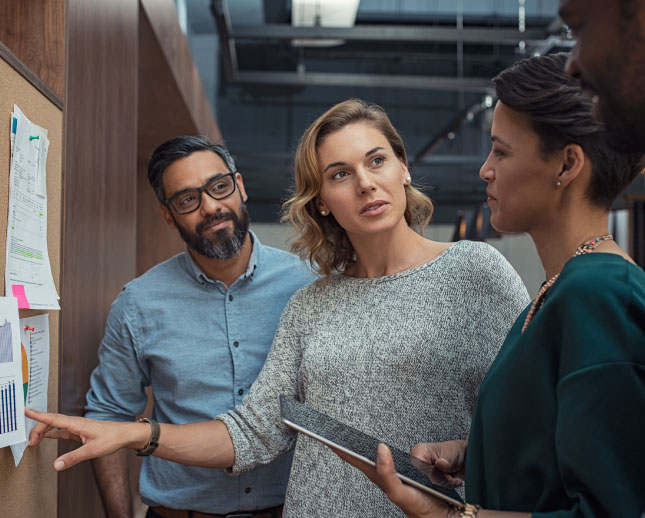 A group of office workers gathered around a corkboard with various sheets of paper pined to it.